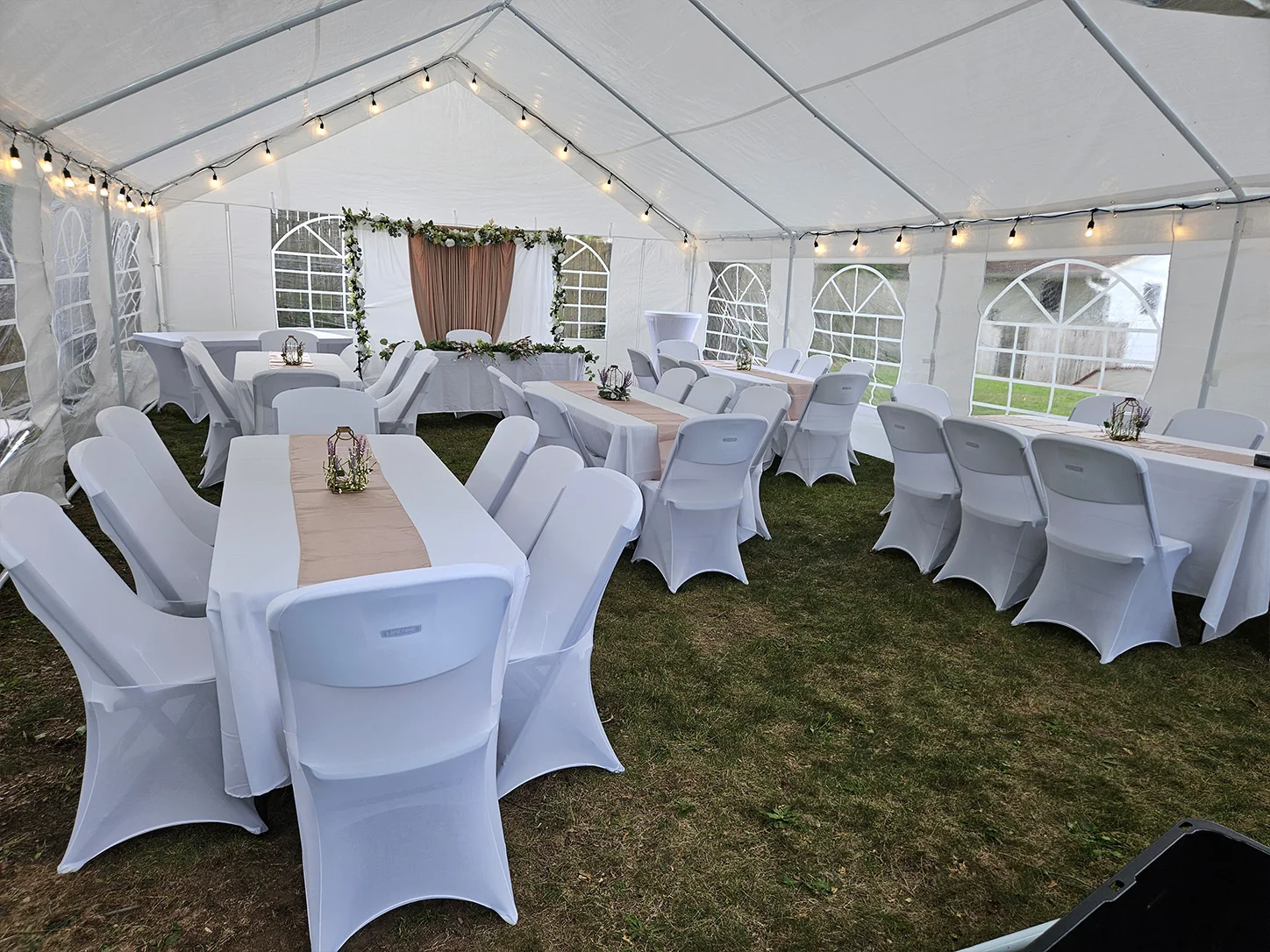 Styled white event tent with covered tables, chairs, bistro lights, and floral decor