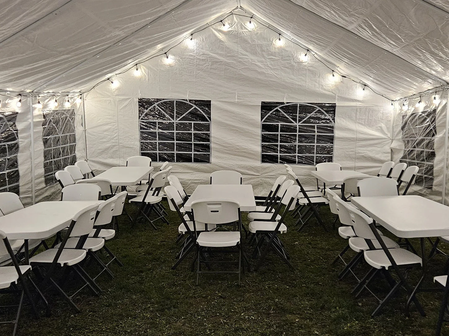 White event tent arranged with tables, folding chairs, sidewalls, and bistro lights